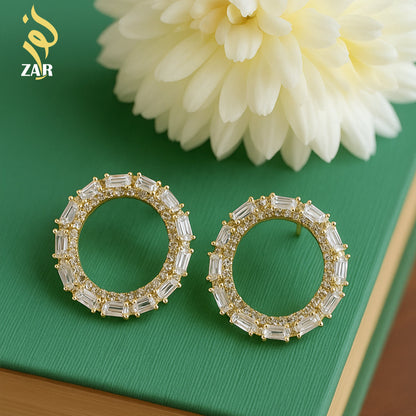 Gold hoop earrings with clear stones on a green surface with a white flower.
