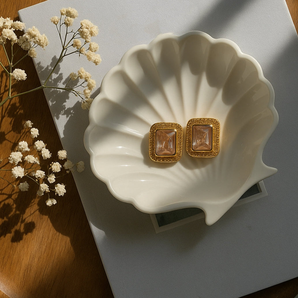 Gold earrings on a white shell-shaped dish with a wooden surface and white flowers in the background.