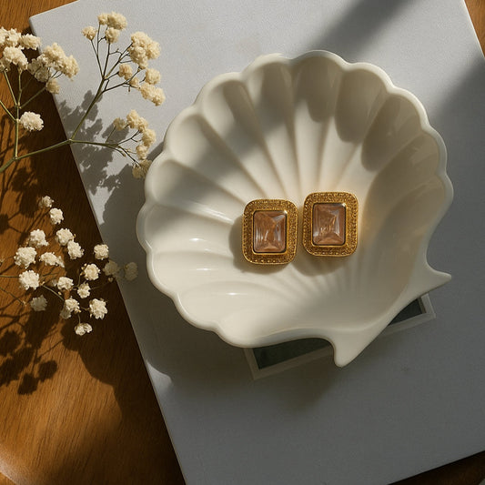 Gold earrings on a white shell-shaped dish with a wooden surface and white flowers in the background.