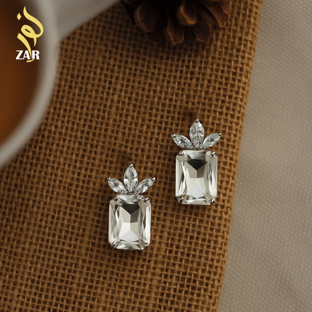 Pair of silver earrings with clear gemstones on a textured brown surface, brand 'ZAR' visible.