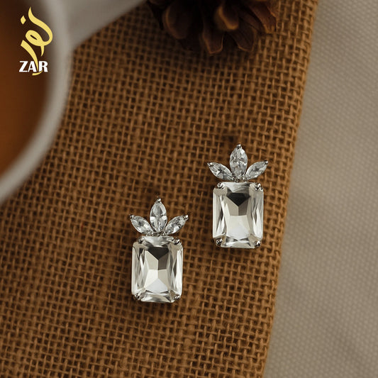 Pair of silver earrings with clear gemstones on a textured brown surface, brand 'ZAR' visible.