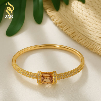 Gold bracelet with a central gemstone on a light background, featuring the brand 'ZAR'.