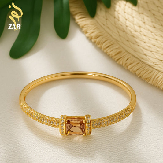 Gold bracelet with a central gemstone on a light background, featuring the brand 'ZAR'.