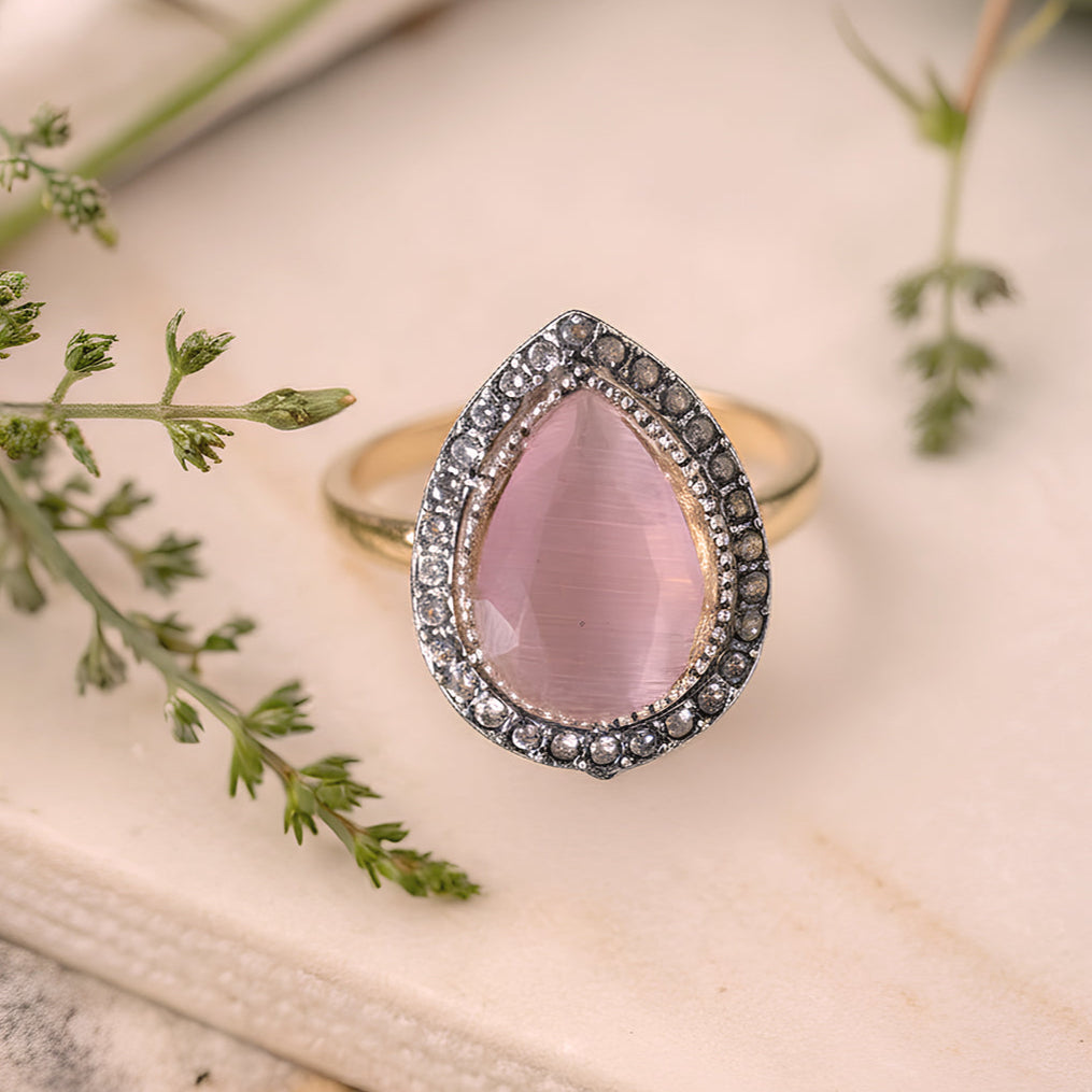 Gold ring with a pink teardrop-shaped gemstone on a light background with greenery